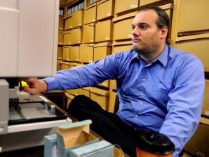 A man in a blue shirt sits down at a computer with boxes behind him